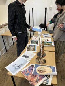 students stand around a table covered with printed artwork images and a small object, talking and pointing while planning a group curatorial layout.