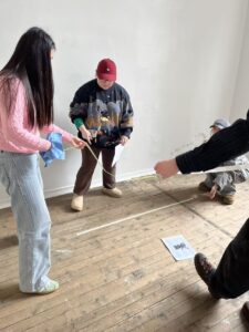  Several students stand and crouch on a wooden floor, holding a tape measure across a taped outline while discussing the dimensions.