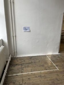 A white wall with pipes on the left; a small printed image is taped to the wall, and a rectangular area is marked on the wooden floor with tape.