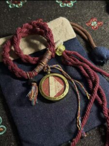 A close-up photograph of a red braided Daoist amulet from Taiqing Palace, Qingdao, placed on a dark blue fabric pouch, with multicoloured threads and a round pendant at the centre.