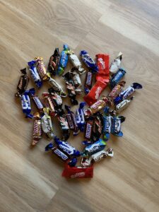 A floor-based installation composed of multiple small, individually wrapped chocolates arranged into a shell-like shape. The candies form an organic, clustered structure on a wooden floor, with different coloured wrappers (red, blue, gold, black, and white) creating visual variation. The arrangement resembles a fragmented shell or organic form, suggesting both accumulation and dispersal.