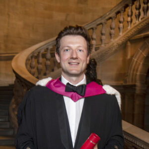 Graduate Calum MacDonald in robes, with a staircase in the background. 