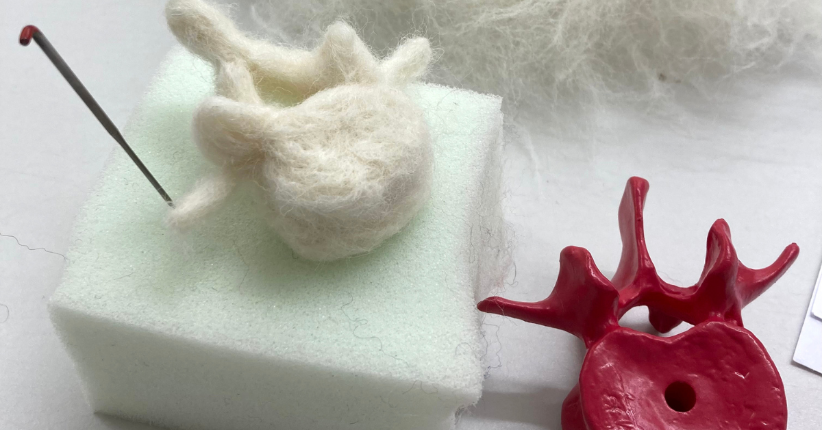 A white felted vertebra sits on a felting mat with a felting needle stuck into it. To its right is a red plastic cast of a vertebra, which is acting as the model for the felted version.