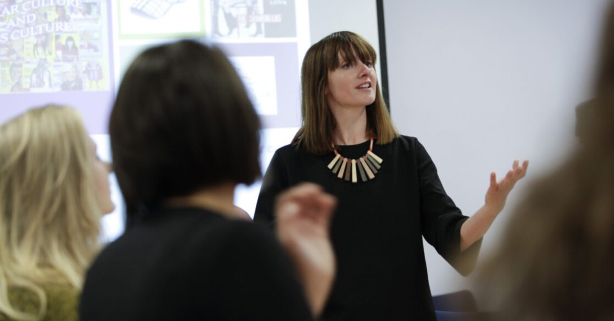 A female academic teaches a class of students, she gestures with her hands