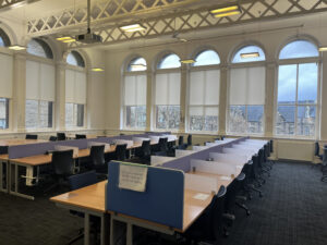 Rows of computer desk and chairs sit in a large airy room. Along the walls are large arched windows, trees and and another building are glimpsed through them.