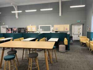 Brightly coloured chairs stools sit beside tables and dark green booths in a large, airy room.