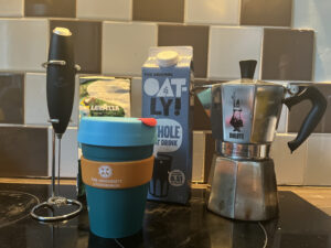 A milk frother, bag of coffee, University of Edinburgh keep cup and a coffee pot stand in a line on a kitchen worktop.