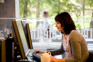 A woman sits at a desk using a desktop computer in a cafe with large windows, with a teapot and cup in front of her.