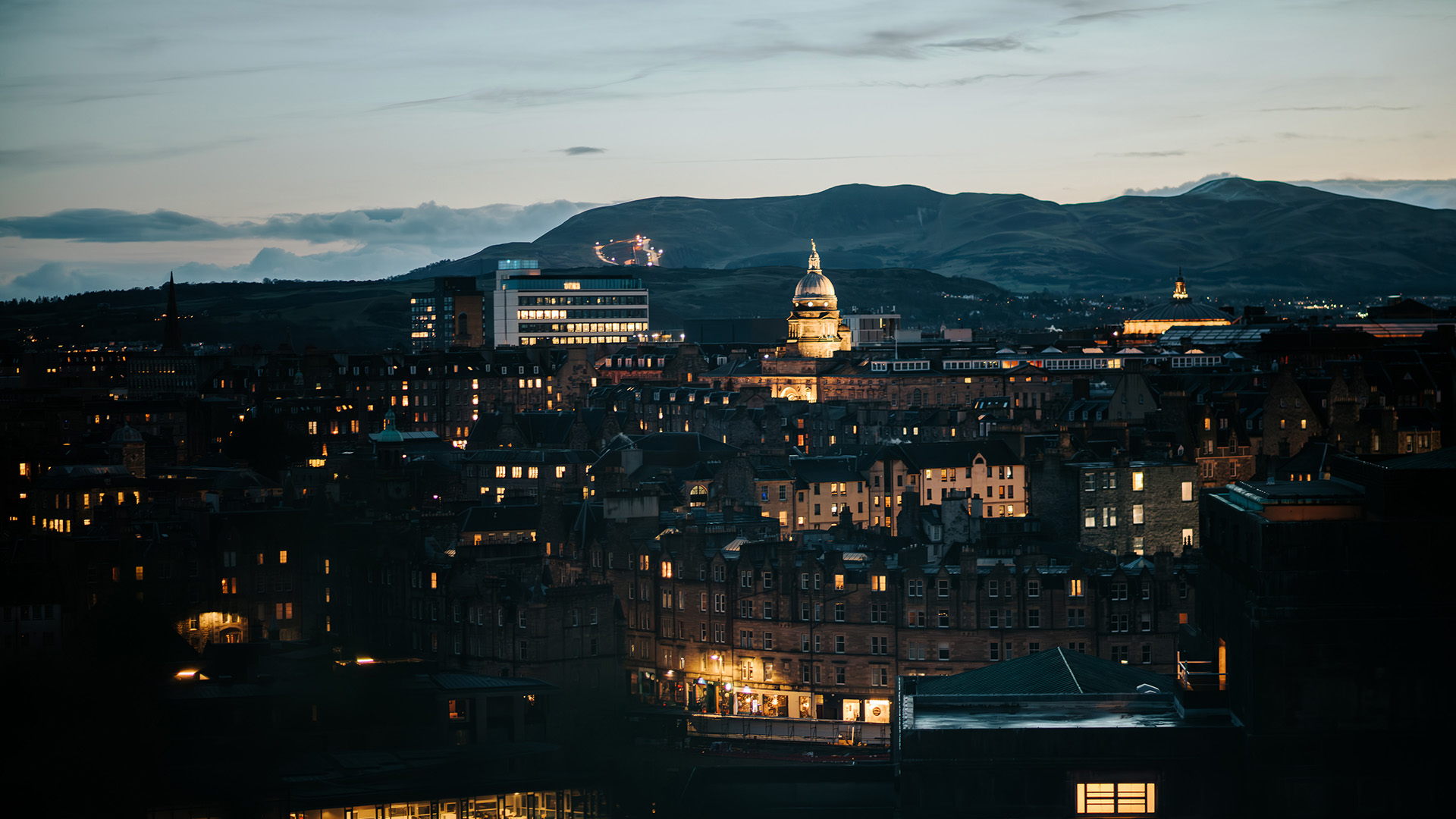 Cityscape of Edinburgh at dusk - with illuminated buildings, distant hills, and a cloudy sky in the background.