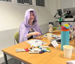 Rachel at a session for GEAS, Edinburgh University Roleplaying Society. She wears a lilac coloured cloak and laughs at the camera