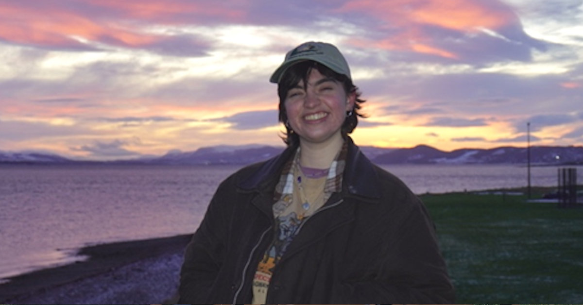 Rachel stands against a sunset, smiling broadly at the camera. She has dark hair and wears a baseball cap