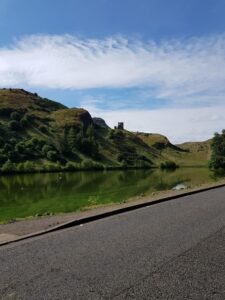 St Margaret's Loch in Holyrood Park. The road passes the Loch where you can see a ruined building ona hill beside the water under a clear blue sky.