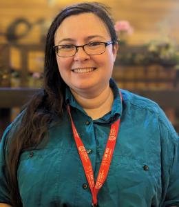 Dr Alice Wolff wears glasses and a teal shirt with a red lanyard. She smiles at the camera, standing indoors with blurred shelves and flowers in the background.