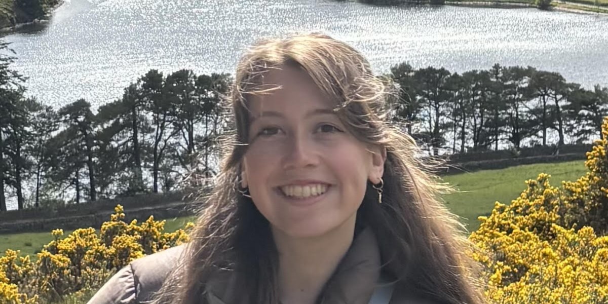 Alice has long fair hair, and smiles directly at the camera against a background of trees and water beyond