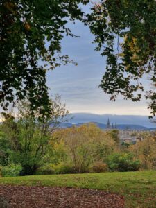 A scenic view across the Royal Botanic Garden Edinburgh, known as The Botanics. Lush green trees frame distant city spires under a cloudy blue sky, conveying tranquility and natural beauty.
