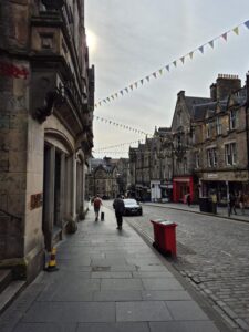 Cockburn Street in the historic Old Town of Edinburgh. Old stone buildings, colorful bunting overhead, and people strolling on the pavement.