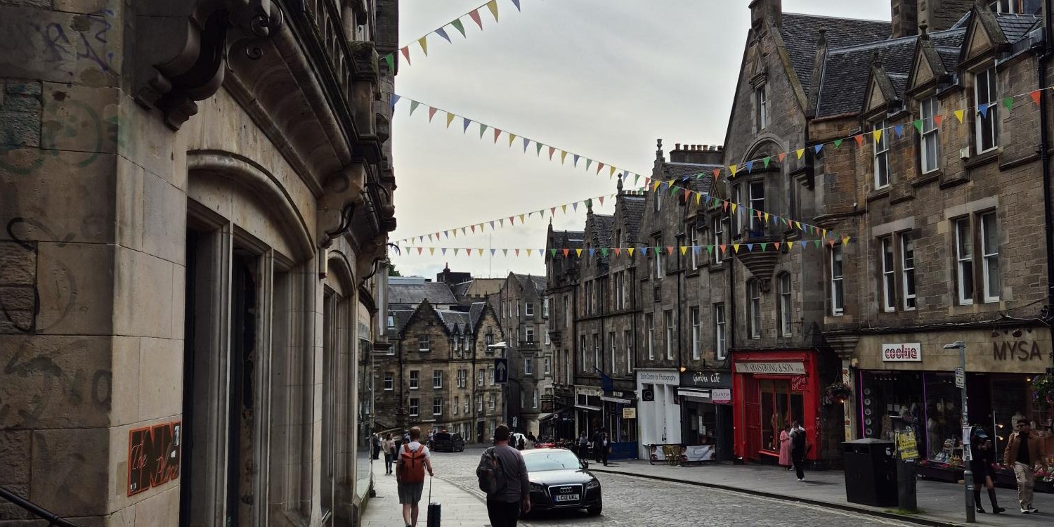 Cockburn Street in the historic Old Town of Edinburgh. Old stone buildings, colorful bunting overhead, and people strolling on the pavement.