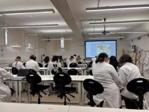 A group of students in white lab coats sit at desks facing a large projection screen in a bright laboratory classroom, focused on a colorful slide.