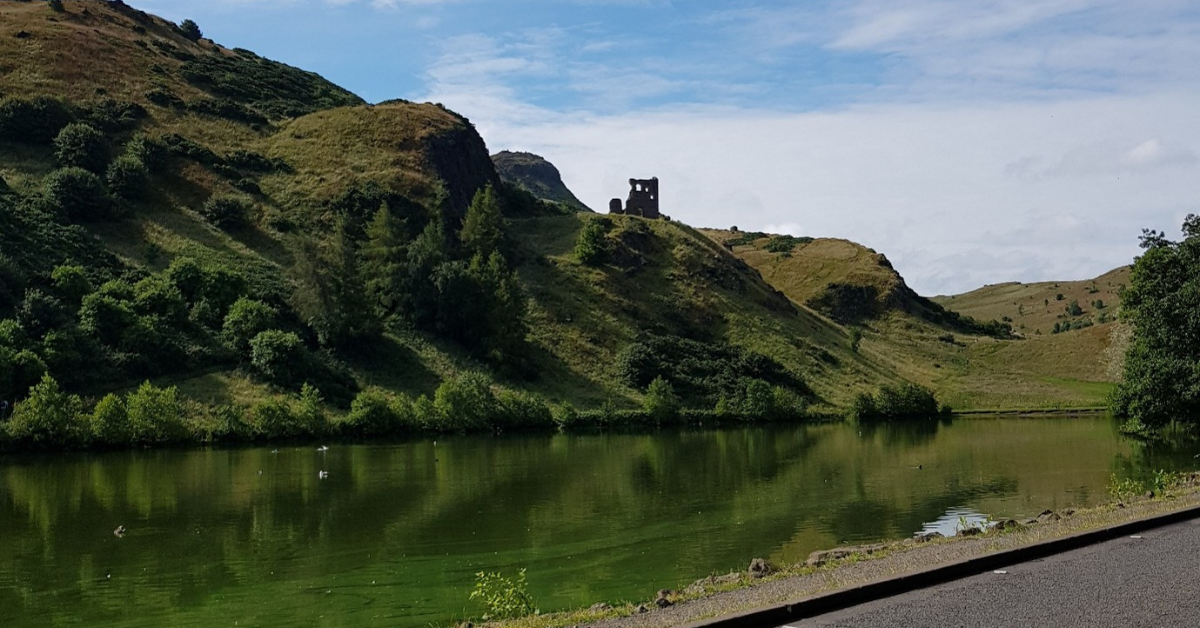 St Margaret's Loch in Holyrood Park. The road passes the Loch where you can see a ruined building ona hill beside the water under a clear blue sky.