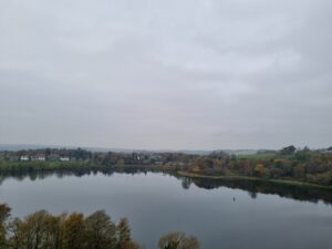 Calm lake under a gray sky, surrounded by trees and houses in the distance. 