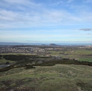 View of Edinburgh in the distance, taken from the Pentland hills. The foreground is lush green hills and the sea is in the background, behind the city skyline.
