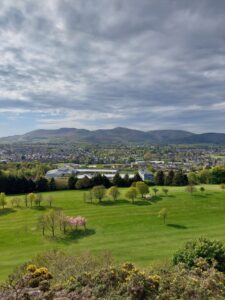 A panoramic view across green grass and tress to rolling hills from an elevated vantage point.