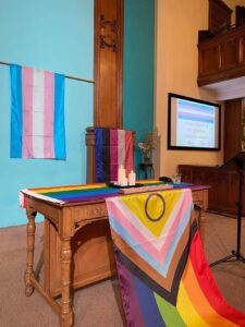 Church altar decorated with trans pride flag hanging on the wall behind the alter with progress pride flag (including intersex circle) draped over the alter.