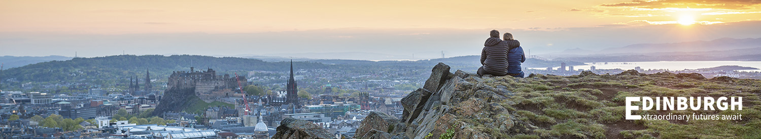 A couple sit on Arthur's Seat with a view of Edinburgh at dusk.