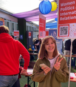 Student Lauren smiles to camera and does the peace sign while stood at a Women in Politics and International Relations stand at the university society fair.