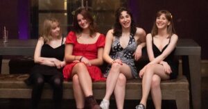 Student Lauren is dressed in a black dress and sits on a bench with three friends as they smile to camera.