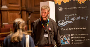 A member of Chaplaincy staff stands behind a table at the Student Information Fair talking to a new student. There is a pull up banner next to the member of staff with The Chaplaincy service details on it. 