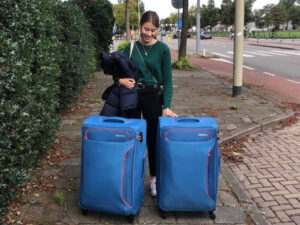 Eva stands on the street with two large blue suitcases in front of her.