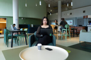 A student sits in the foreground on a comfy chair using a laptop with a phone and coffee cup in a table. Three other students are sat in the background working on laptops of using their phone. The room is decorated with calming shades of green.