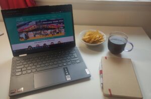 Student Carl's desk with laptop, bowl of crisps a glass of drink and a notebook.