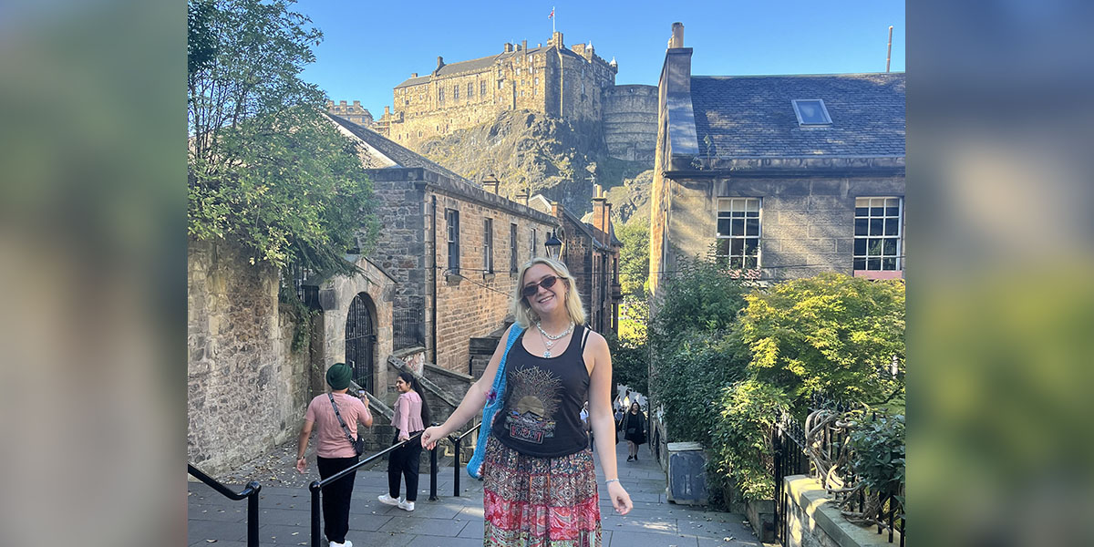 Maisie stands at the top of some steps. Behind her in the sunshine is Edinburgh Castle.