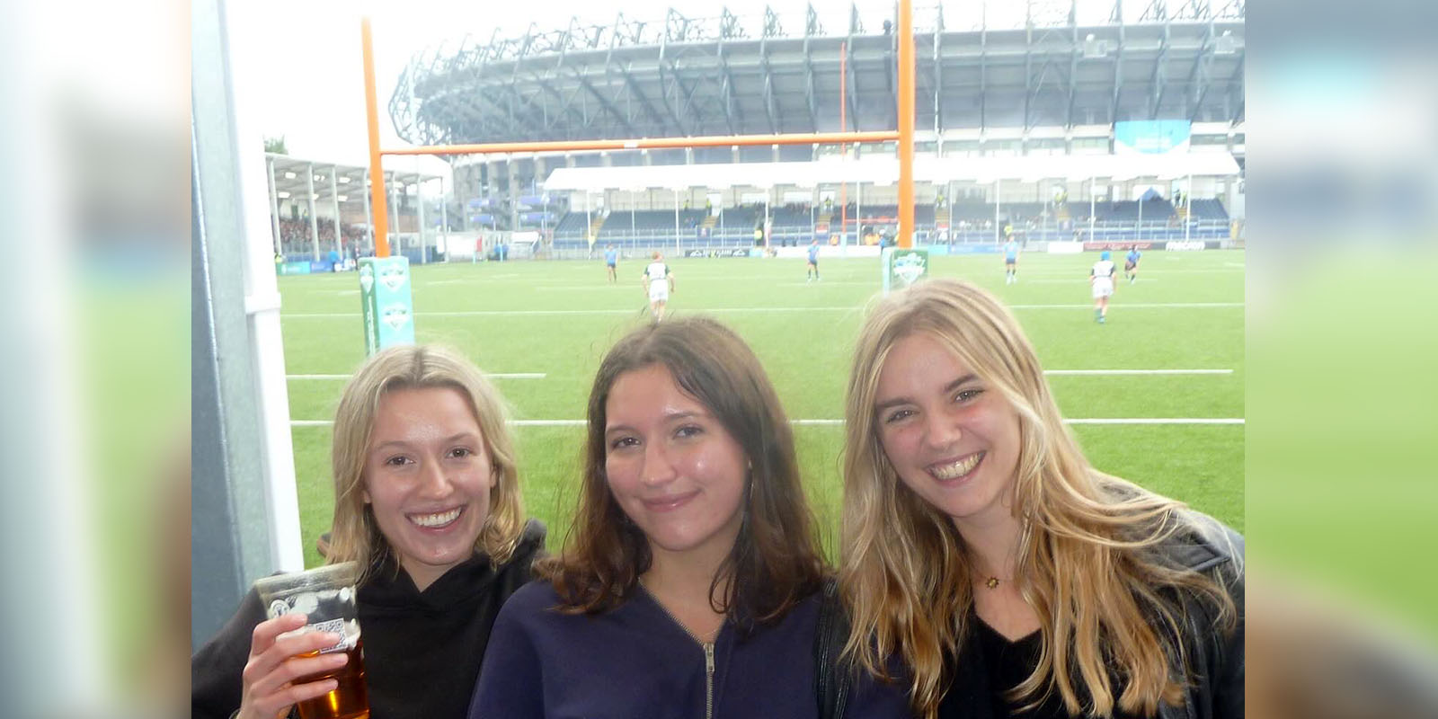 Annabel and two friends standing in front of a rugby game.