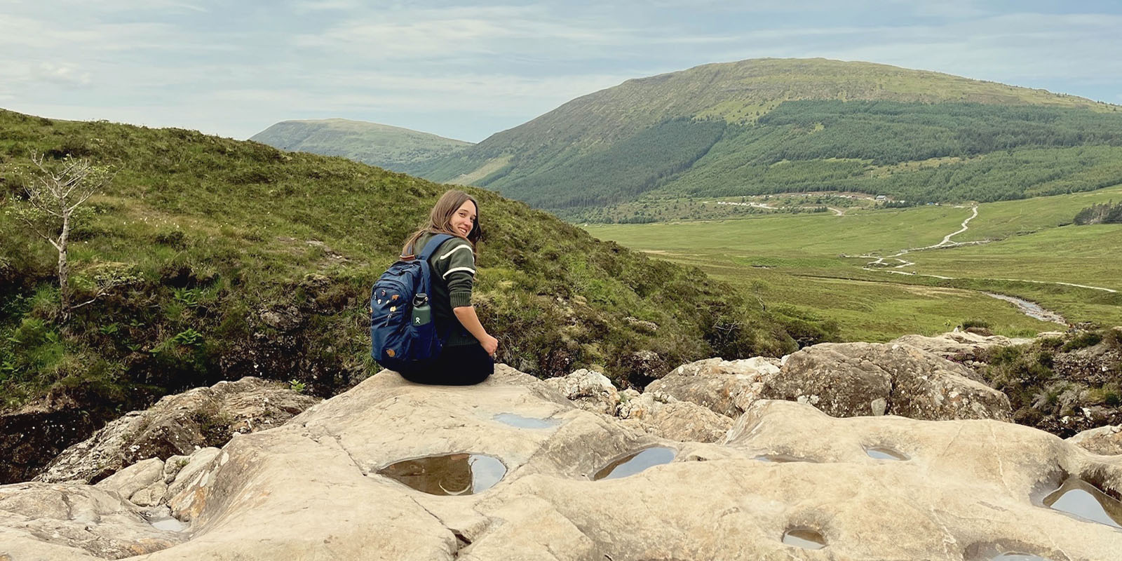 Bella sits in front a a beautiful Scottish landscape, looking back and smiling.
