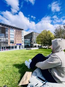 A student sits with their hood up on the grass at the Quad at King's Buildings. Several university buildings are in the background and there's a cloudy blue sky.