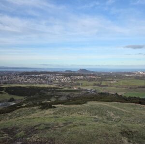 View of Edinburgh in the distance, taken from the Pentland hills. The foreground is lush green hills and the sea is in the background, behind the cityline.