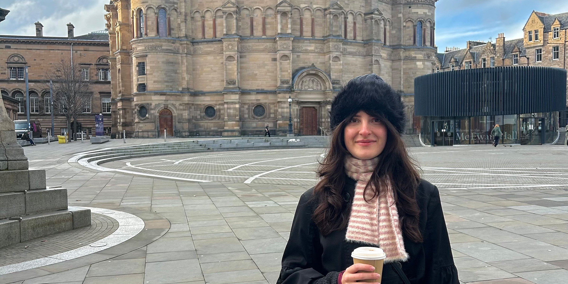 Student, Barbara, stands in front of McEwan Hall holding a takeaway cup. She wears a dark, furry hat and a white and red scarf.