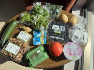 A selection of groceries sit on top of a table, including food such as blueberries, sardines, potatoes and eggs.