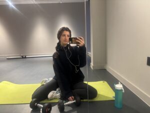 Barbara sits on a yoga mat, with two dumbells, in front of a mirror and takes a selfie.