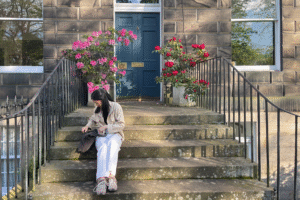 A person sat looking through their bag on steps leading to a front door. Two plants with colourful flowers are seen by the door.