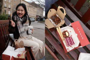 A person smiling for a photograph; pastries and desserts in boxes on a park bench.