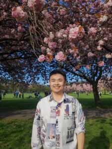 Student, Ian, stands and smiles in front of a pink blossom tree in full bloom.