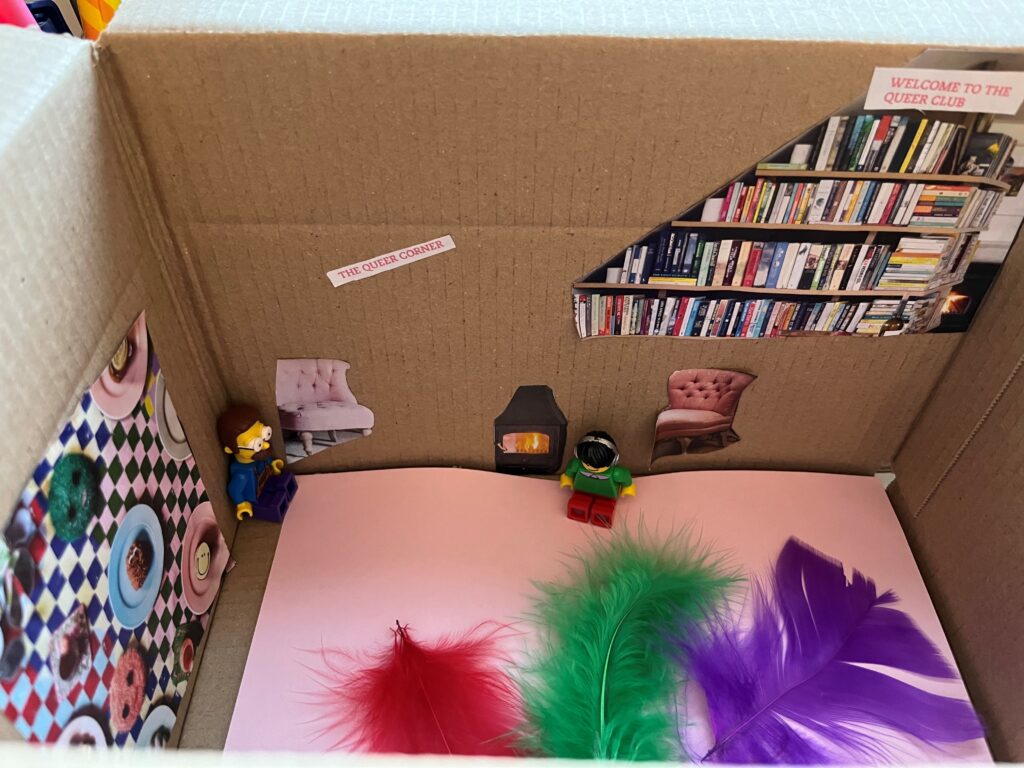 A craft box with two lego people, a lot of feather and a sign saying 'queer corner'