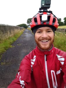 Andrew cycling to work, wearing red helmet and jacket on country road
