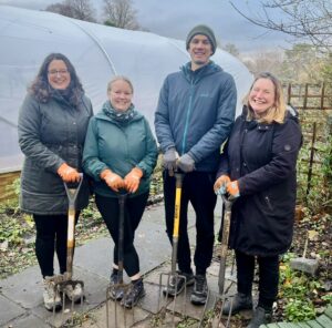 Three women and a man holding garden forks, standing in front of a polytunnel. The weather is wet and they are wearing raincoats.
