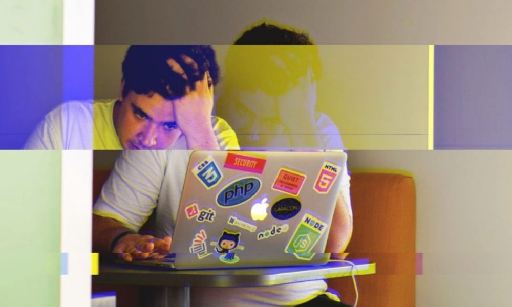 Photograph of man seated at a table showing frustration while working on a laptop