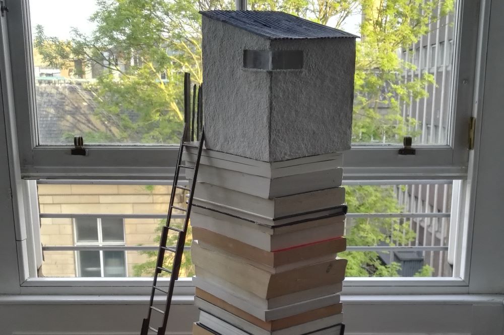 Photograph of a sculpture made by the author featuring a model treehouse located on the top of a stack of books with a small model stepladder resting against the house.